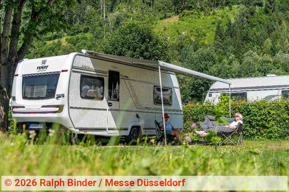 Wohnwagen auf einem Campingplatz inmitten einer grünen Berglandschaft. Unter der ausgefahrenen Markise sitzen zwei Personen auf Campingstühlen und genießen das sonnige Wetter. | Foto: Ralph Binder / Messe Düsseldorf