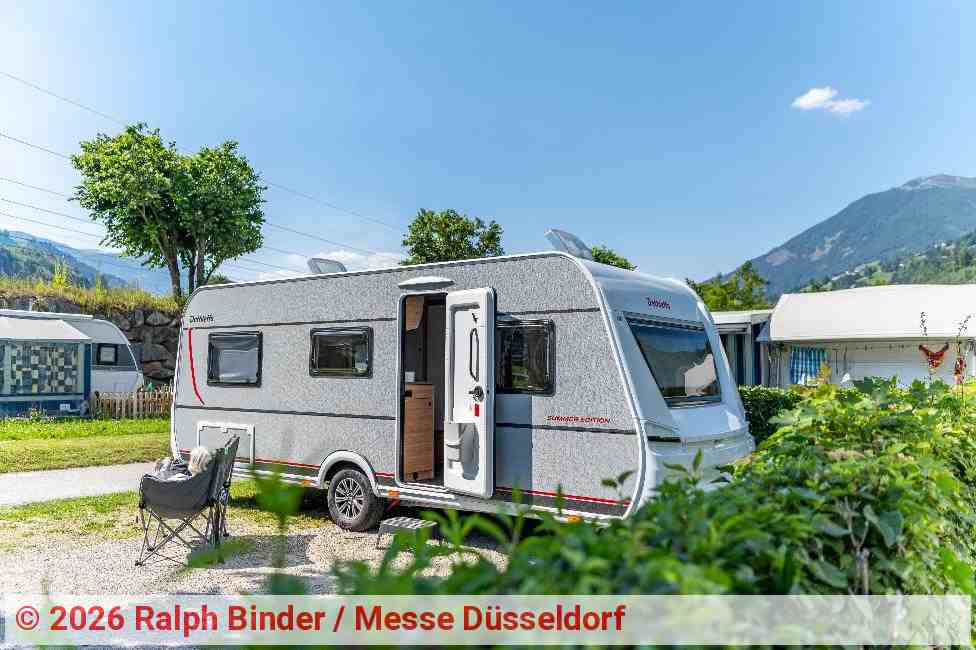 A modern caravan at a campsite in Tyrol, set against an alpine mountain backdrop. The caravan door is open, with a camping chair in front of it. In the background, further camping accommodation, green trees and the wooded slopes of the Alps can be seen beneath a blue sky. | Photo: Ralph Binder / Messe Düsseldorf