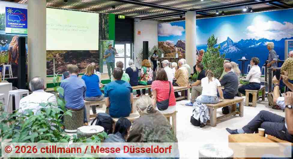 Vortrag auf einer Messebühne beim CARAVAN SALON in Düsseldorf. Zahlreiche Besucher sitzen auf Holzbänken und verfolgen eine Präsentation zum Thema Outdoor- und Caravaning-Tourismus. Die Veranstaltungsfläche ist mit Pflanzen, Bäumen und großformatigen Naturmotiven gestaltet. | Foto: ctillmann / Messe Düsseldorf